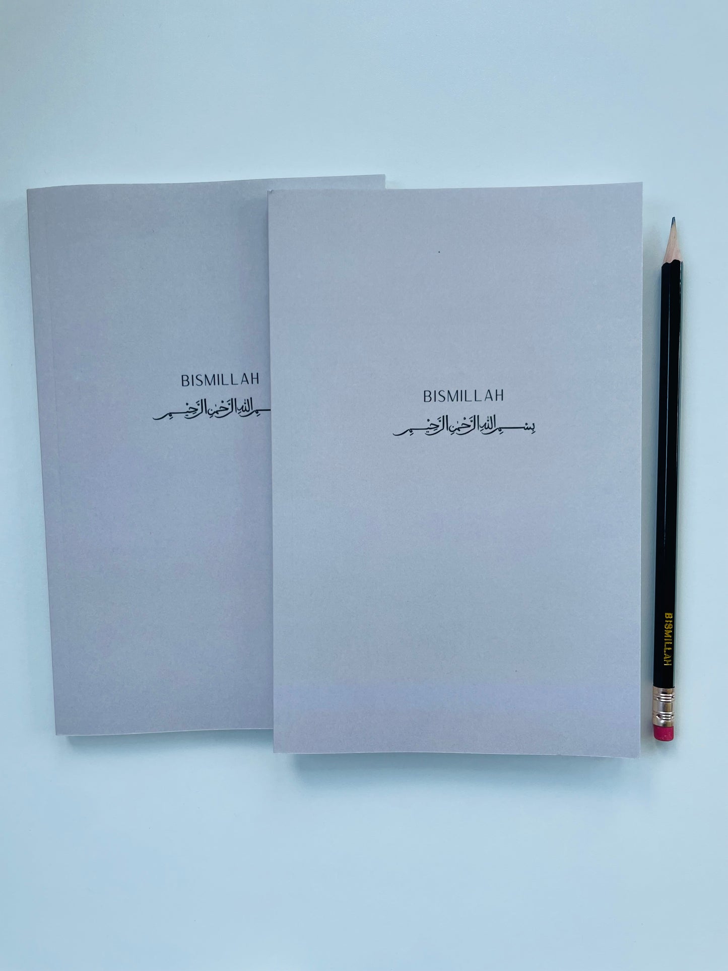 Two Grey notebooks with 'Bismillah' text on a light blue background, accompanied by a pencil.