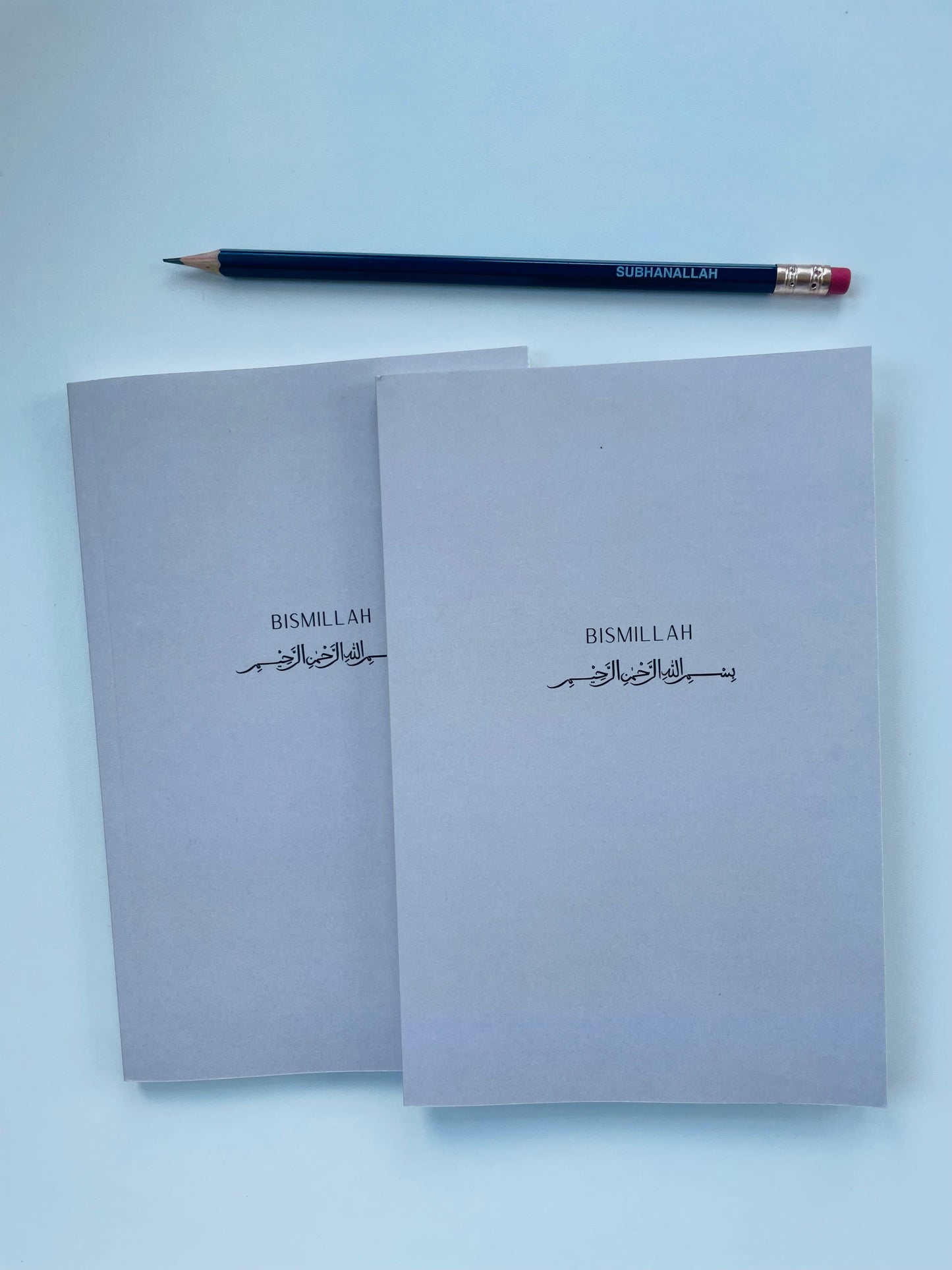 Two grey notebooks with 'Bismillah' text on a light blue background, accompanied by a pencil.